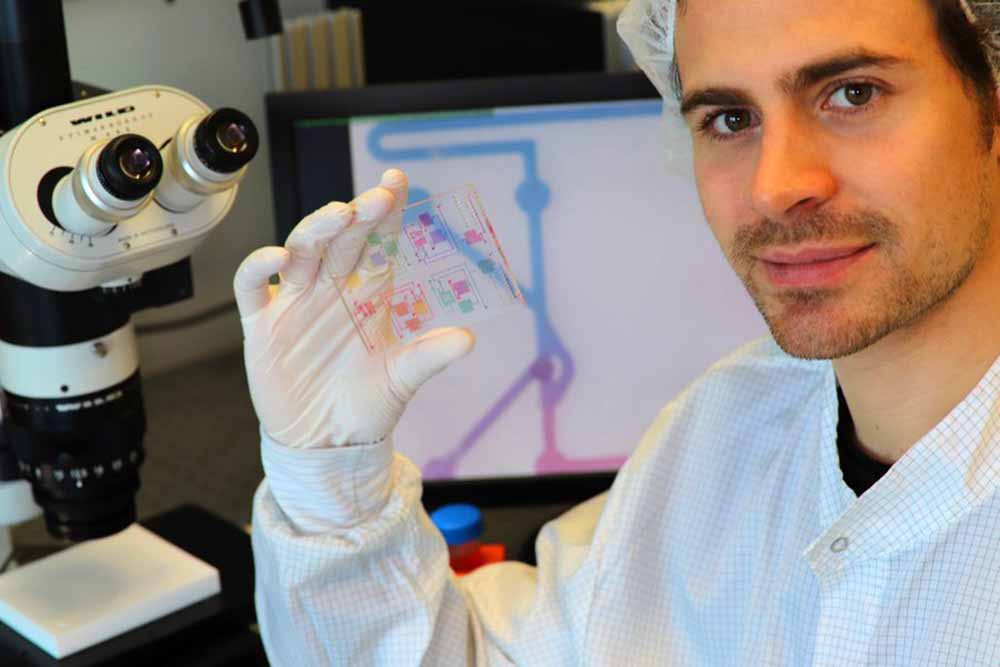 Doktorand Anthony Beck, Mit-Entwickler der Technologie, hält einen chemischen Schaltkreis mit gefärbten Analysemedien. Auf dem Monitor im Hintergrund ist das Mikroskopiebild eines der chemischen Transistoren zu sehen. Foto: TU Dresden