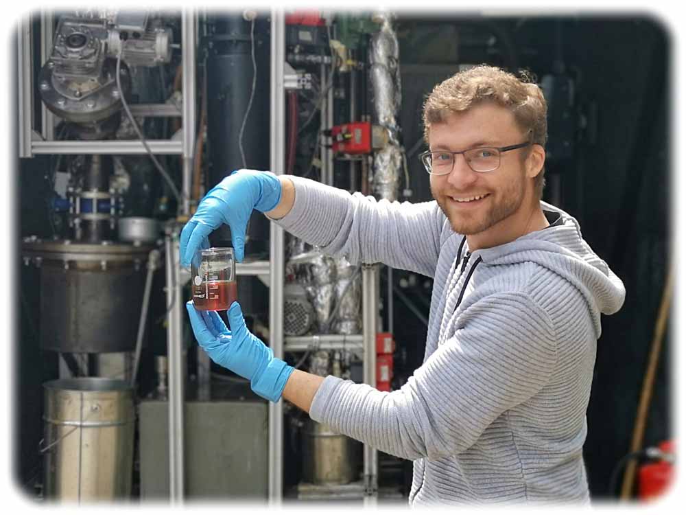 Biofabrik-Projektleiter und Ingenieur Paul Foth zeigt ein Messglas mit einem Kraftstoff-Gemisch, das der Pyrolyse-Container im Hintergrund aus Plastemüll erzeugt hat. Foto: Heiko Weckbrodt