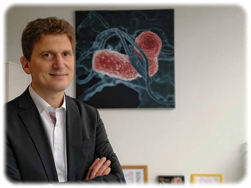 Professor Alexander Dalpke leitet das Institut für medizinische Mikrobiologie und Hygiene im Uniklinikum Dresden. Die Grafik im Hintergrund zeigt, wie die Tentakel einer Fresszelle ein Bakterium umschlingen. Foto: Heiko Weckbrodt