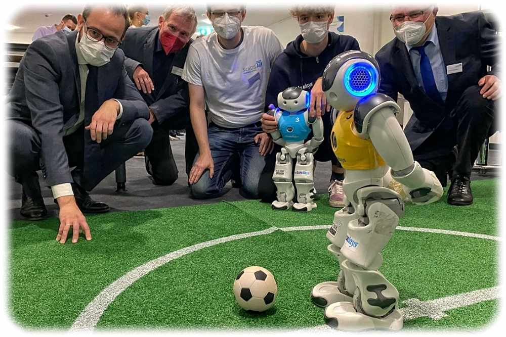 Wissenschaftsminister Sebastian Gemkow (links) schaut gemeinsam mit jungen Besuchern des "Living Labs" in Leipzig zu, wie sich Nao-Roboter mit KI-Unterstützung als Fußballer verdingen. Foto: SMWK