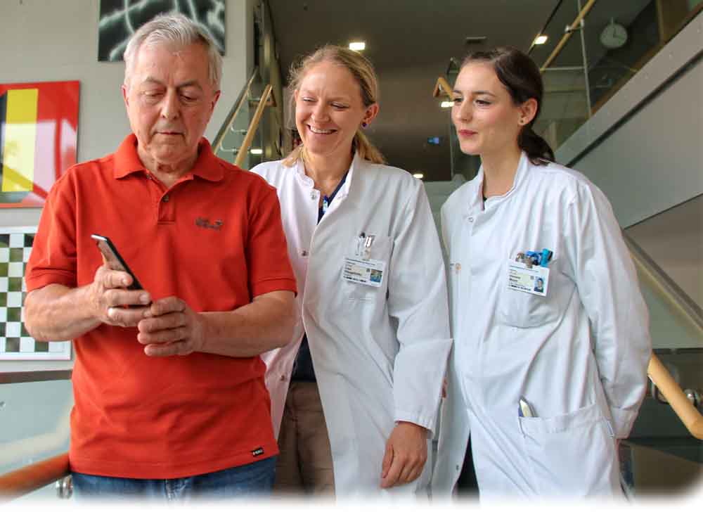 Patient Horst Maier in der Uniklinik Dresden sein Smartphone, auf dem er die Parkinson-Früherkennungs-App für Studienzwecke installiert hat, Die Neurologen Dr. Lisa Klingelhöfer und Dr. Simone Mayer schauen ihm zu. Foto: Heiko Weckbrodt